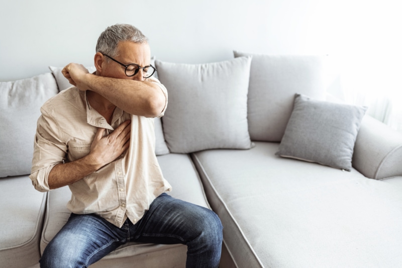 How to Spot a Cracked Heat Exchanger. Man coughs in his elbow while sitting on sofa at home.