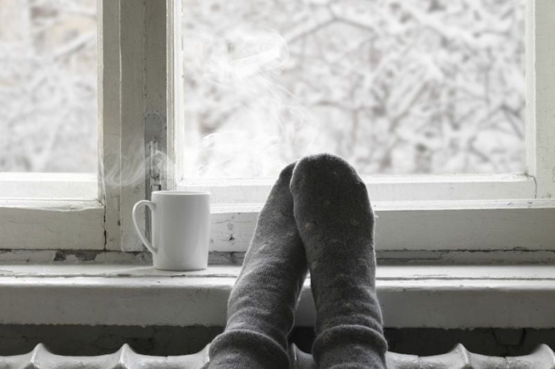 How Do I Keep My Heat During Extreme Cold Weather? Image shows person's socked feet resting on windowsill next to a hot drink.